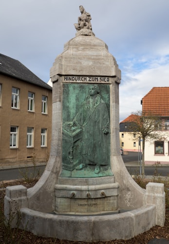 A historical monument featuring a sculpted relief of a person in traditional robes, possibly a notable figure, with inscriptions in German. The monument appears to be made of stone and bronze, with a smaller statue atop it depicting a figure in a contemplative pose. Surrounding the monument are buildings and a clear sky with some clouds.