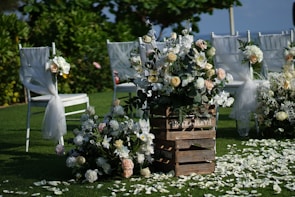 An outdoor setting with an arrangement of white chairs decorated with sheer fabric and floral bouquets. A wooden crate labeled 'FARM FRESH' supports a mix of white and pink flowers. Petals are scattered across the grass, enhancing the serene and romantic atmosphere.