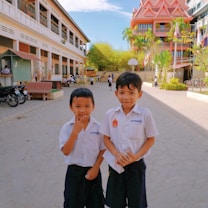 Two young boys in school uniforms are standing in a spacious school courtyard with a modern building in the background. Other students can be seen in the backdrop, along with trees, a basketball hoop, and parked bicycles.
