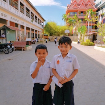 Two young boys in school uniforms are standing in a spacious school courtyard with a modern building in the background. Other students can be seen in the backdrop, along with trees, a basketball hoop, and parked bicycles.
