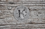 A metallic coin featuring the word 'KARBO' at the top, with 'DECENTRALIZED EXCHANGE MEDIUM' inscribed around the edge. The center shows a large letter 'K' over a circuit board pattern, placed on a textured wooden surface with visible grain and an aged appearance.