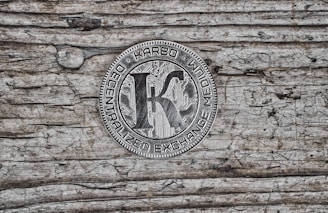 A metallic coin featuring the word 'KARBO' at the top, with 'DECENTRALIZED EXCHANGE MEDIUM' inscribed around the edge. The center shows a large letter 'K' over a circuit board pattern, placed on a textured wooden surface with visible grain and an aged appearance.
