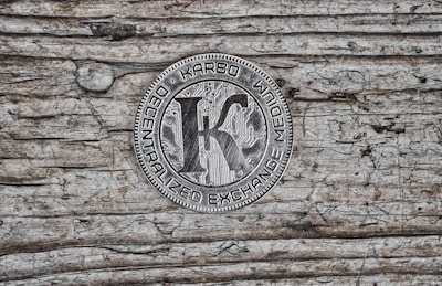 A metallic coin featuring the word 'KARBO' at the top, with 'DECENTRALIZED EXCHANGE MEDIUM' inscribed around the edge. The center shows a large letter 'K' over a circuit board pattern, placed on a textured wooden surface with visible grain and an aged appearance.