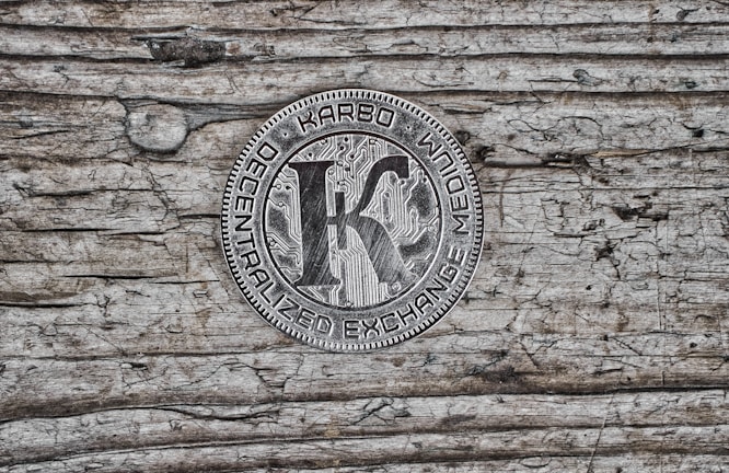 A metallic coin featuring the word 'KARBO' at the top, with 'DECENTRALIZED EXCHANGE MEDIUM' inscribed around the edge. The center shows a large letter 'K' over a circuit board pattern, placed on a textured wooden surface with visible grain and an aged appearance.