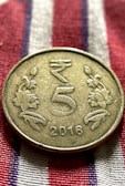 A metallic coin is placed on a fabric with red, white, and blue stripes. The coin has an embossed design featuring the denomination '5' in the center, flanked by floral motifs. The year '2016' is inscribed at the bottom.