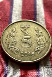 A metallic coin is placed on a fabric with red, white, and blue stripes. The coin has an embossed design featuring the denomination '5' in the center, flanked by floral motifs. The year '2016' is inscribed at the bottom.