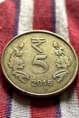 A metallic coin is placed on a fabric with red, white, and blue stripes. The coin has an embossed design featuring the denomination '5' in the center, flanked by floral motifs. The year '2016' is inscribed at the bottom.