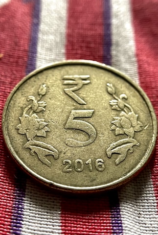 A metallic coin is placed on a fabric with red, white, and blue stripes. The coin has an embossed design featuring the denomination '5' in the center, flanked by floral motifs. The year '2016' is inscribed at the bottom.