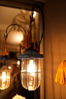 A warm, glowing light bulb encased in a metal and glass lantern, adorned with a decorative tassel, casting a soft illumination that highlights the surrounding area in a cozy ambiance.