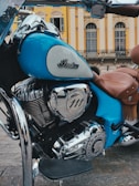 A vintage-style motorcycle with a polished chrome engine and a distinctive emblem. The bike is predominantly blue with brown leather seats, set against an elegant building with classic architecture and arched windows.