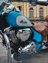 A vintage-style motorcycle with a polished chrome engine and a distinctive emblem. The bike is predominantly blue with brown leather seats, set against an elegant building with classic architecture and arched windows.