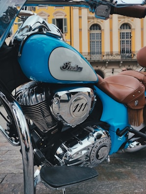 A vintage-style motorcycle with a polished chrome engine and a distinctive emblem. The bike is predominantly blue with brown leather seats, set against an elegant building with classic architecture and arched windows.