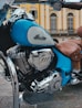 A vintage-style motorcycle with a polished chrome engine and a distinctive emblem. The bike is predominantly blue with brown leather seats, set against an elegant building with classic architecture and arched windows.