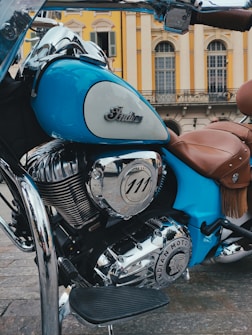 A vintage-style motorcycle with a polished chrome engine and a distinctive emblem. The bike is predominantly blue with brown leather seats, set against an elegant building with classic architecture and arched windows.