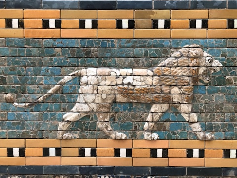 A decorative wall relief features a striding lion rendered in vibrant glazed bricks. The lion is depicted in profile, showcasing textured details in the fur and mane. The surrounding bricks display a pattern of blue and teal hues, interspersed with geometric designs in yellow and black.