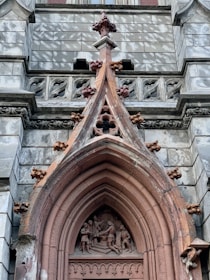 A detailed architectural feature from a historical building, showcasing a sculpted scene with figures within a pointed arch. The stone structure includes intricate carvings, ornamental elements, and weathered textures. The design has gothic influences, evident in the pointed arch and decorative elements around it.