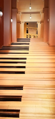 A staircase designed to resemble a piano keyboard, with black and white steps. The space is well-lit with hanging lights and ornate sconces. The walls have an elegant architectural design with arches and columns.