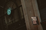 An exterior view of a building features a prominently displayed turquoise clock with Roman numerals, mounted on an ornate metal framework. Next to it, a neon sign reading 'JAZZ BAR' is affixed to the stone wall, giving a vintage and urban feel.