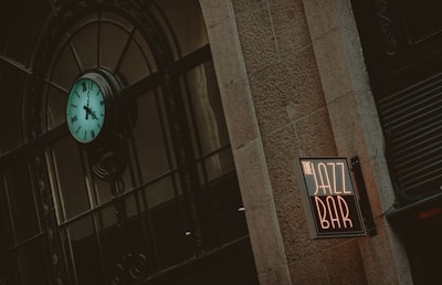 An exterior view of a building features a prominently displayed turquoise clock with Roman numerals, mounted on an ornate metal framework. Next to it, a neon sign reading 'JAZZ BAR' is affixed to the stone wall, giving a vintage and urban feel.
