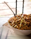 A bowl filled with stir-fried noodles topped with various vegetables like cabbage and possibly peppers. The noodles appear to be well-seasoned, and there is a fork placed in the bowl. The setting suggests a casual dining environment with natural light streaming in.