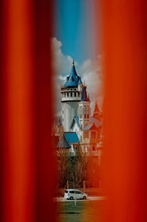 A red sedan parked beside a historic European castle under a bright blue sky