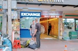 Two men stand outside an exchange booth, with one carrying a backpack. The booth is adjacent to a hotel entrance with the sign 'Sleep With Inn'. A scooter and some travel bags are on the sidewalk next to a red stool. There is a traffic cone nearby and various shop signs in different languages are visible.