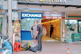Two men stand outside an exchange booth, with one carrying a backpack. The booth is adjacent to a hotel entrance with the sign 'Sleep With Inn'. A scooter and some travel bags are on the sidewalk next to a red stool. There is a traffic cone nearby and various shop signs in different languages are visible.
