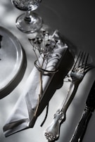 A sophisticated table setting with a crystal glass, a white napkin wrapped with a metal ring and adorned with a dried flower, positioned beside an elegant silver fork and knife on a white surface.
