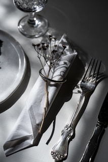A sophisticated table setting with a crystal glass, a white napkin wrapped with a metal ring and adorned with a dried flower, positioned beside an elegant silver fork and knife on a white surface.