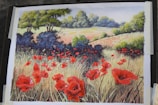 A vibrant painting depicts a scenic landscape with a field full of red poppies. There are patches of tall grasses, lush green trees, and a distant horizon lined with more flowers and foliage. The artwork captures a peaceful and serene rural scene under a clear blue sky.