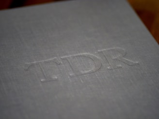 A close-up of a gray textured surface with embossed uppercase letters TDR. The surface has a fabric-like appearance, suggesting it might be the cover of a book or an album.