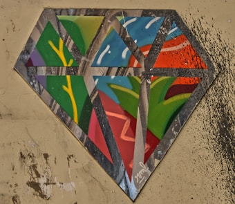 A diamond-shaped piece of street art with a splatter of multicolored paint on a rough, beige wall. The artwork consists of geometric segments filled with vibrant colors including green, red, blue, and yellow, delineated by metallic silver lines.