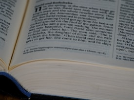 An open book displaying a passage from a religious text. The focus is on a story about David and Bathsheba, with clear, small-sized printed text on white pages. The edges of the pages have a slight shadow, and a blue cloth is partially visible, hinting at the book cover.