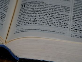 An open book displaying a passage from a religious text. The focus is on a story about David and Bathsheba, with clear, small-sized printed text on white pages. The edges of the pages have a slight shadow, and a blue cloth is partially visible, hinting at the book cover.