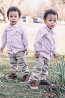Two young children are standing on a grassy area. They are wearing matching outfits consisting of light-colored shirts, khaki pants, and brown shoes. The setting appears to be outdoors on a sunny day with trees and parked cars visible in the background.