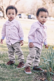 Two young children are standing on a grassy area. They are wearing matching outfits consisting of light-colored shirts, khaki pants, and brown shoes. The setting appears to be outdoors on a sunny day with trees and parked cars visible in the background.