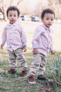 Two young children are standing on a grassy area. They are wearing matching outfits consisting of light-colored shirts, khaki pants, and brown shoes. The setting appears to be outdoors on a sunny day with trees and parked cars visible in the background.