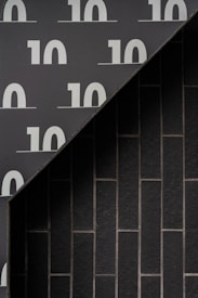 The image features a modern architectural wall design, with a diagonal line dividing the upper left and lower right sections. The upper left section displays large, repeated numeral '10' patterns set against a dark gray background. The lower right section consists of vertically aligned rectangular tiles in a darker shade with subtle grout lines.