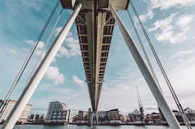 A modern bridge viewed from below, featuring thick supporting cables and a clear sky in the background. Below the bridge, a river is visible along with several contemporary buildings lining the waterfront. A construction crane can be seen to the right among the buildings.
