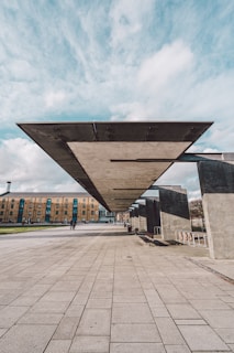 A modern architectural structure featuring a large horizontal concrete beam supported by vertical pillars. The beam appears suspended above a paved open space. In the background, there is a brick building with a series of windows. The sky above is partly cloudy with shades of blue and white.