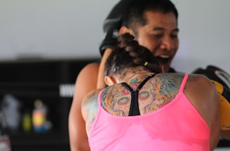 A woman with an intricate back tattoo and braided hair is engaged in a boxing session with a trainer. She is wearing a pink tank top and appears focused on the activity. The trainer, standing in front, is holding up boxing pads and is smiling.
