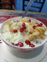 A bowl filled with sliced bananas, red pomegranate seeds, and what appears to be a dairy product like yogurt or cream, all mixed together. A teaspoon is placed in the bowl, which sits on a table with a colorful tablecloth.