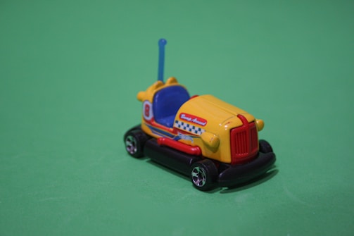 A small, toy bumper car is placed on a plain green background. The car is brightly colored, with a primarily yellow body, blue and red details, and the number '8' on its sides. The toy features a whimsical design with an antenna, decorative patterns, and small wheels.