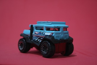 A small, toy vehicle with a rugged design, featuring a predominantly blue body and black, oversized wheels. Decorative elements, such as painted designs and patterns, are visible on the sides. It is positioned against a solid pinkish-red background.