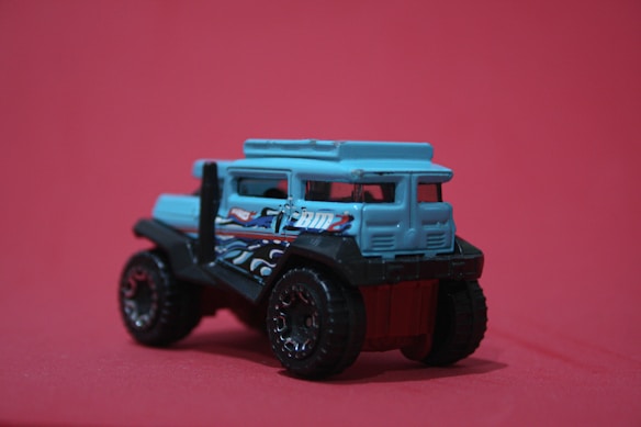 A small, toy vehicle with a rugged design, featuring a predominantly blue body and black, oversized wheels. Decorative elements, such as painted designs and patterns, are visible on the sides. It is positioned against a solid pinkish-red background.