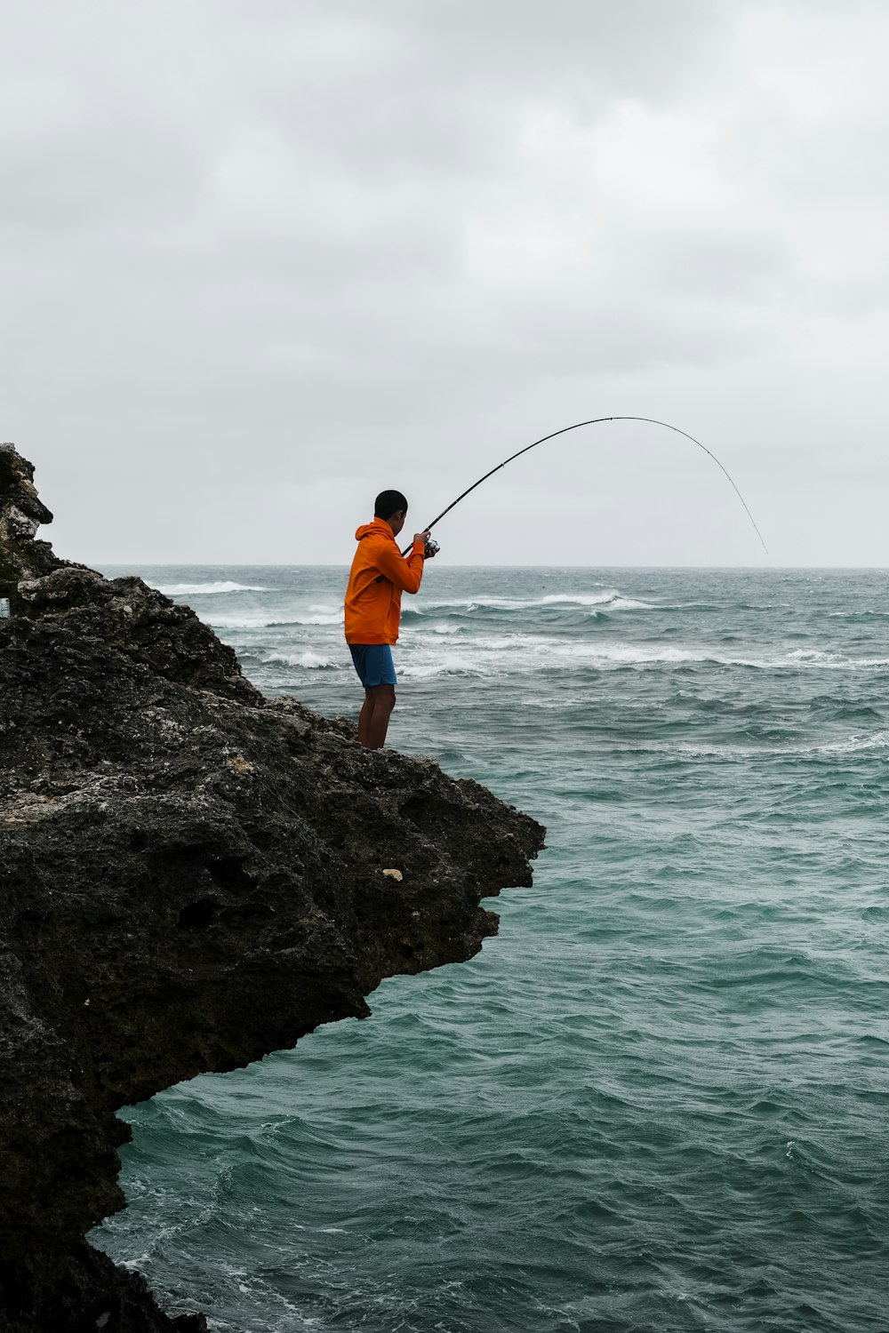 man in orange jacket and blue denim jeans fishing on sea during daytime