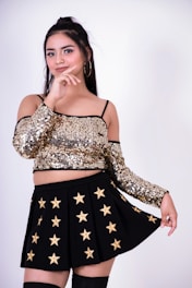 A woman dressed in a glittering gold sequined off-shoulder top and a black skirt adorned with gold stars. She has long dark hair tied in a half-up style and is posing with a confident expression, gently touching her chin.