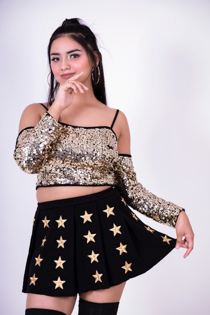 A woman dressed in a glittering gold sequined off-shoulder top and a black skirt adorned with gold stars. She has long dark hair tied in a half-up style and is posing with a confident expression, gently touching her chin.