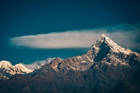 A majestic snow-capped mountain peak under a clear blue sky, with a layer of clouds hovering just above the lower slopes. The sunlight illuminates parts of the mountain, highlighting its rugged texture and creating a sense of grandeur.