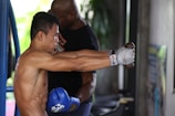 A person engaging in boxing training, wearing boxing gloves and hand wraps, with a coach or trainer standing nearby for assistance. The environment appears to be a gym with equipment visible, including a boxing ring.
